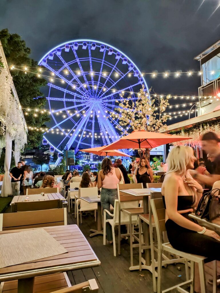 A group of people sitting at tables and chairs in front of a ferris wheel