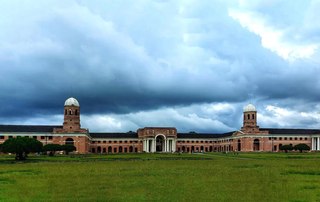 A large brick building called Forest Research Institute Dehradun with a large lawn and a cloudy sky