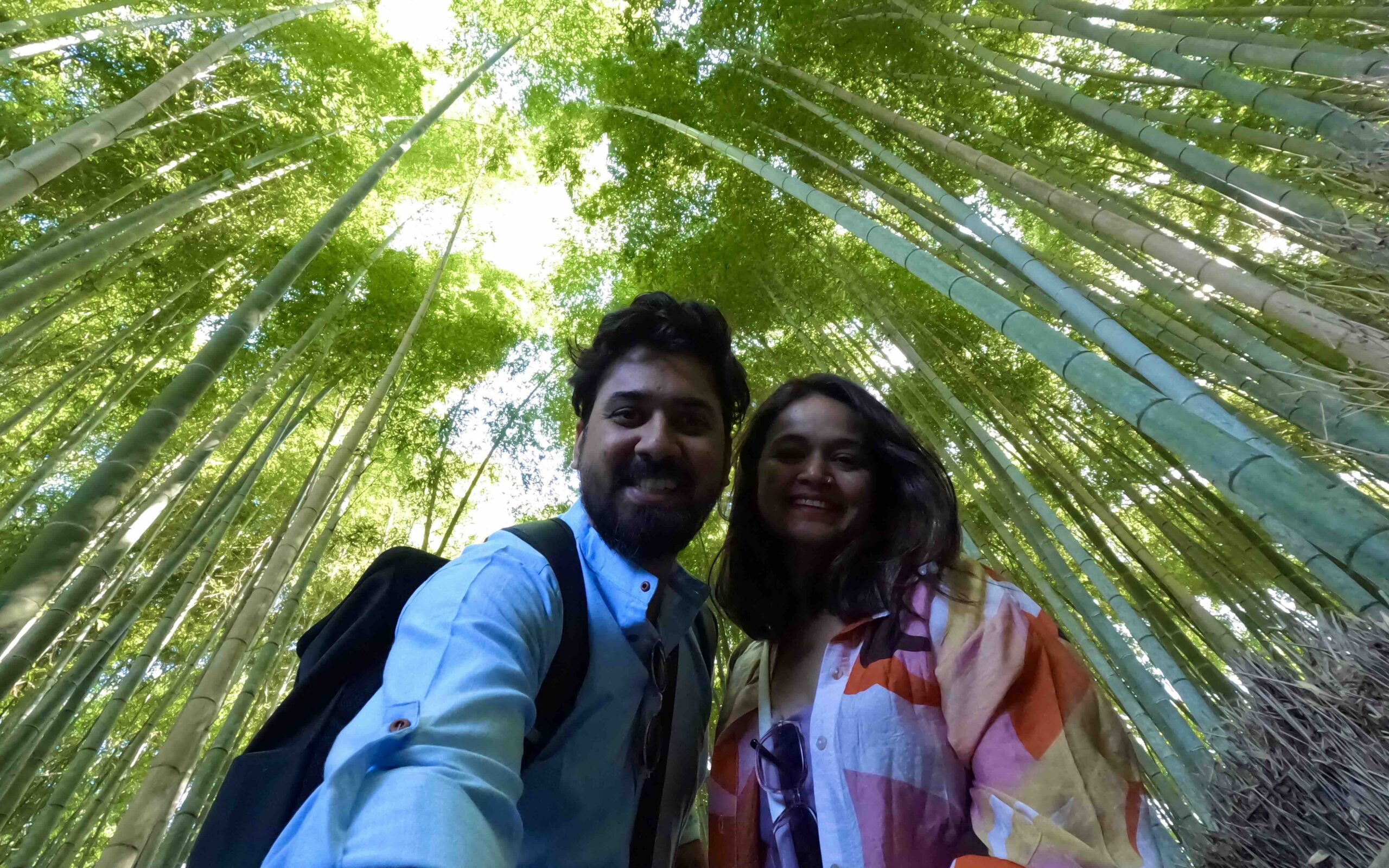 A man and a woman taking a selfie. In the background is the Adashino Bamboo Forest in Japan. The trees are green and long.
