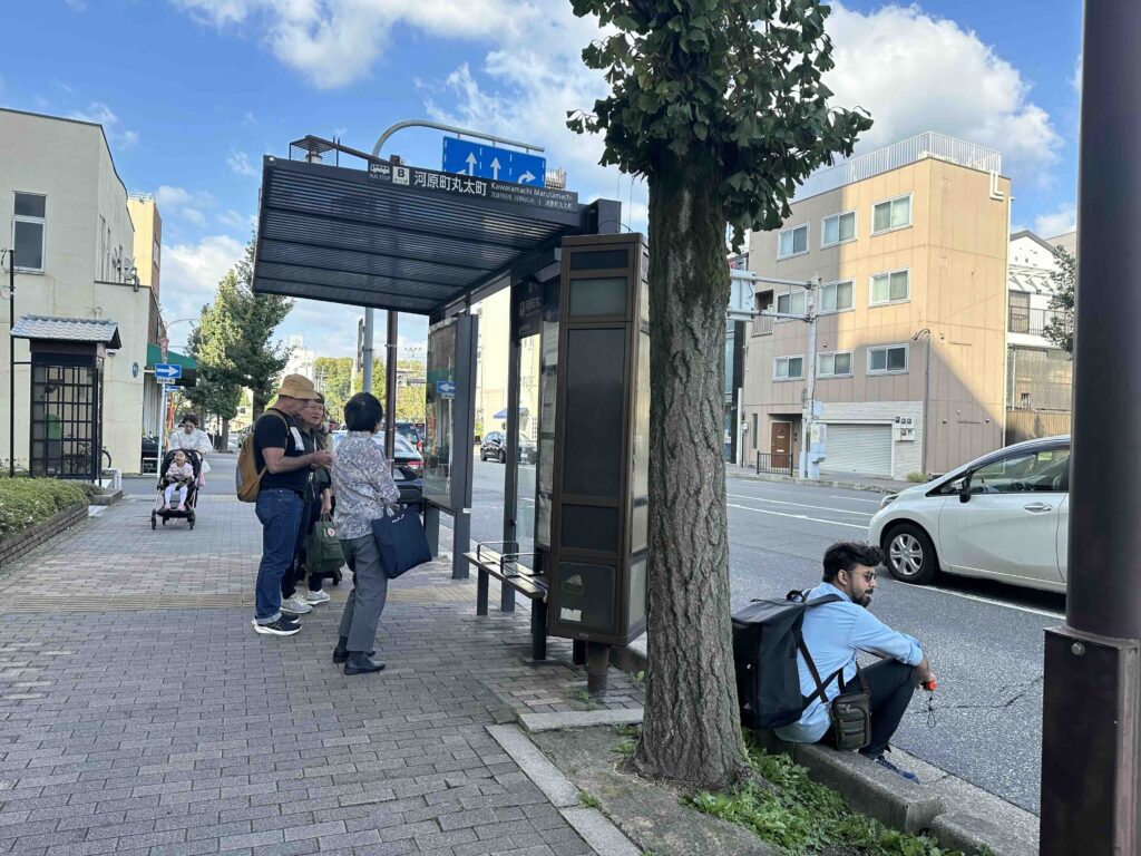 A person sitting at the right corner of a bus stop. 2 people are at the bus stop standing. On the way to Adashino Bamboo Forest.