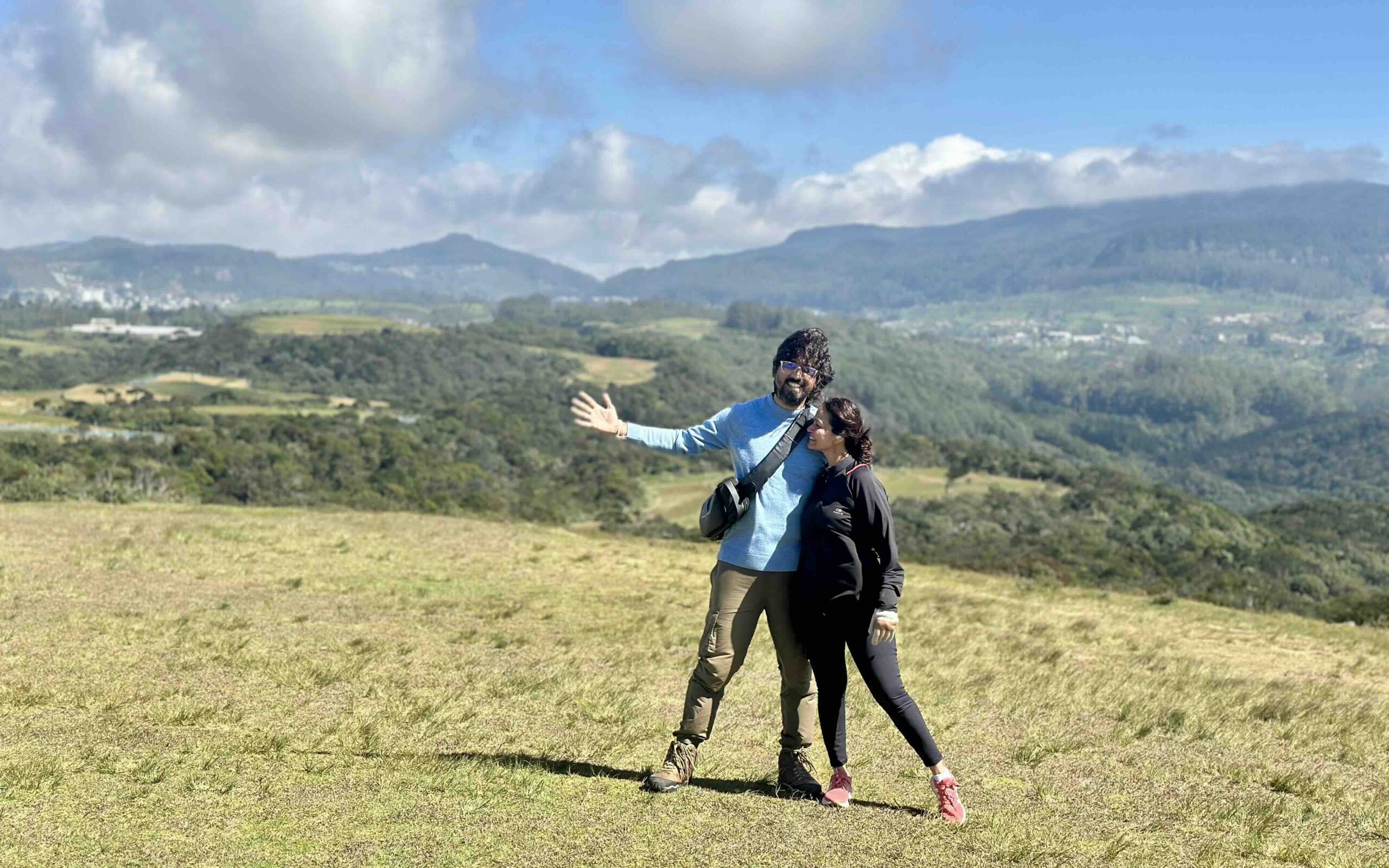 A man in a blue sweater and a woman in black clothes standing in an open grassland with mountains behind at Moon Plains, Nuwara Eliya Sri Lanka